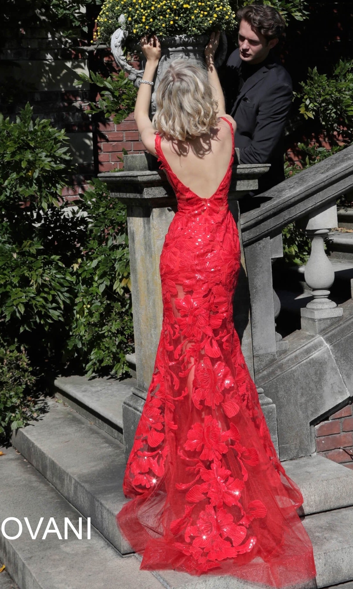 A woman wears a sleeveless red Jovani Embroidered Long Prom Dress with Sequins featuring an open back and floral embroidery, standing with a man on stone steps.
